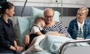 Teenager comforting elderly patient in hospital bed, surrounded by family members, illustrating emotional support during grief and loss.
