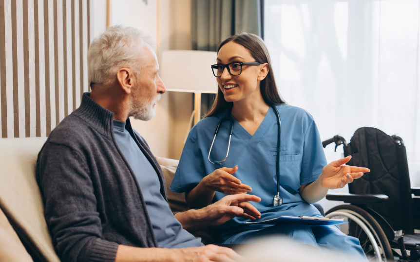 A female nurse in blue scrubs talking with a senior male veteran about hospice care options in a home setting, with a wheelchair visible in the background.