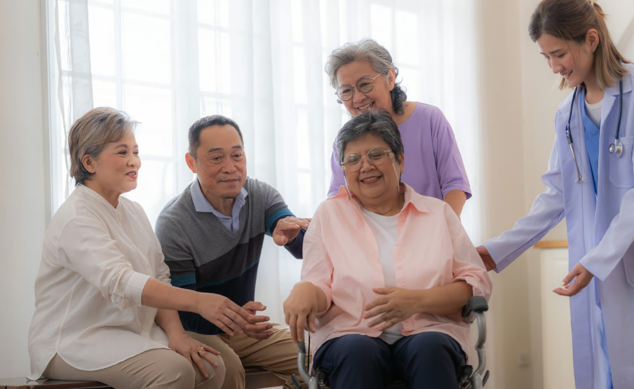 A smiling senior Asian woman sits in a wheelchair surrounded by her supportive family and a medical professional in a bright room.