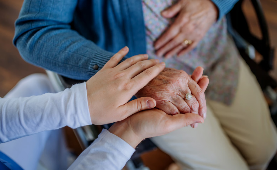 Close-up of a healthcare worker's hands gently holding and comforting an elderly resident's hand in a wheelchair.