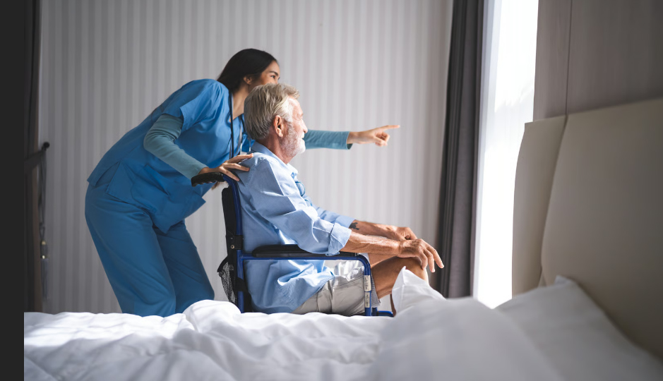 A nurse in blue scrubs stands behind an elderly man in a wheelchair, pointing out a bright window to show him the view from his room.