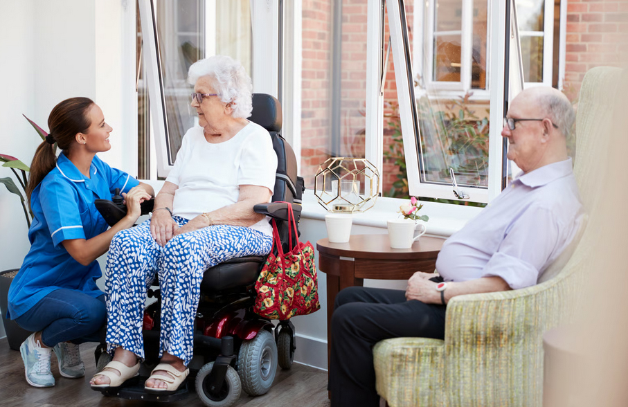 A female hospice nurse in a bright blue uniform smiling and speaking with an elderly woman in a motorized wheelchair alongside a senior man in an armchair near a window.
