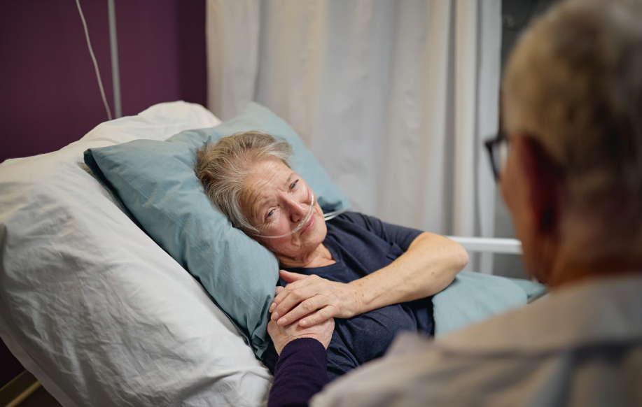 An elderly woman wearing a nasal oxygen cannula lies in a hospital bed, looking up with a gentle smile while a loved one or healthcare worker holds her hand supportively.