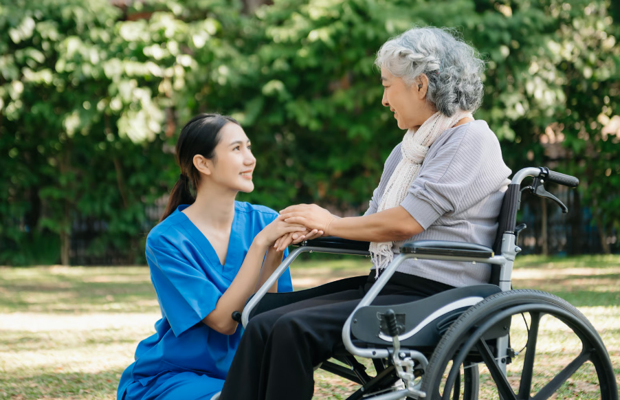 A young female nurse in blue scrubs kneels down on the grass to hold the hands of an elderly Asian woman with gray hair who is sitting in a wheelchair in a sunlit park.