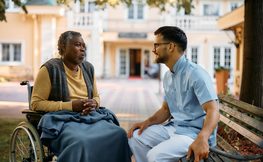 A male hospice caregiver in light blue scrubs sitting on an outdoor bench and speaking compassionately with an elderly Black man in a wheelchair, illustrating the emotional care and counseling support provided to families experiencing anticipatory grief.