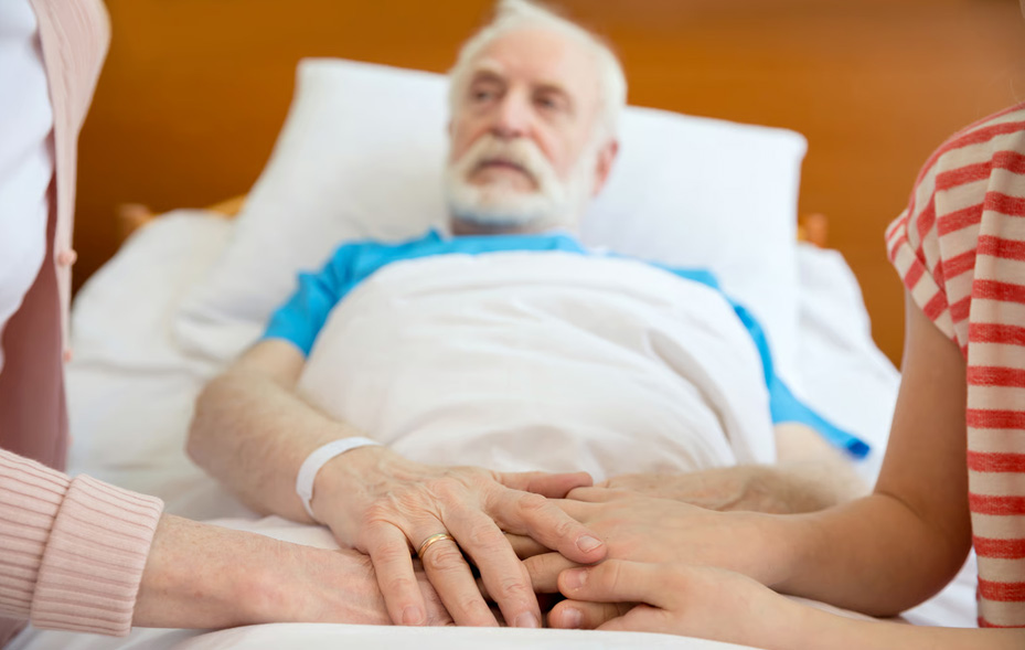 Family members gently holding the hands of an elderly man with a white beard lying in a hospice bed to offer support during his final weeks.