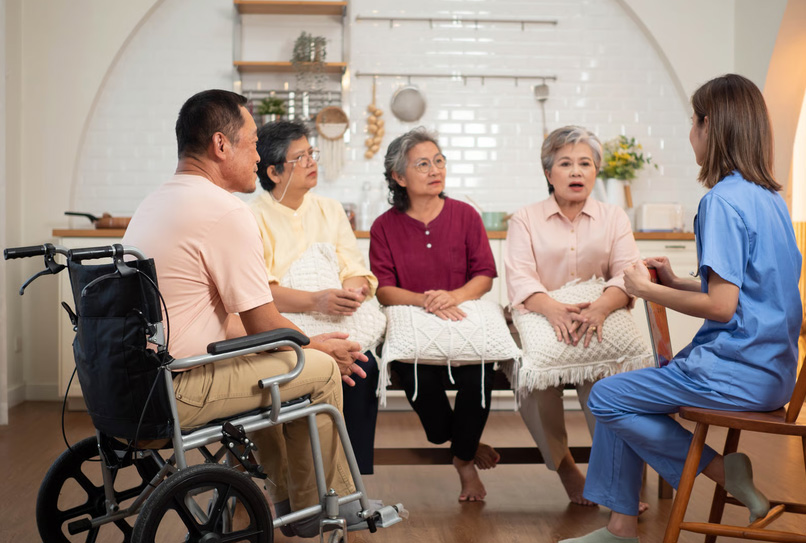 A hospice nurse in blue scrubs discussing care options with a group of elderly family members, including a senior man in a wheelchair, in a home setting.