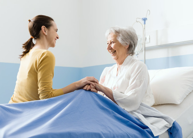 A loving caregiver holding hands and sharing a warm smile with an elderly woman resting in a care bed.