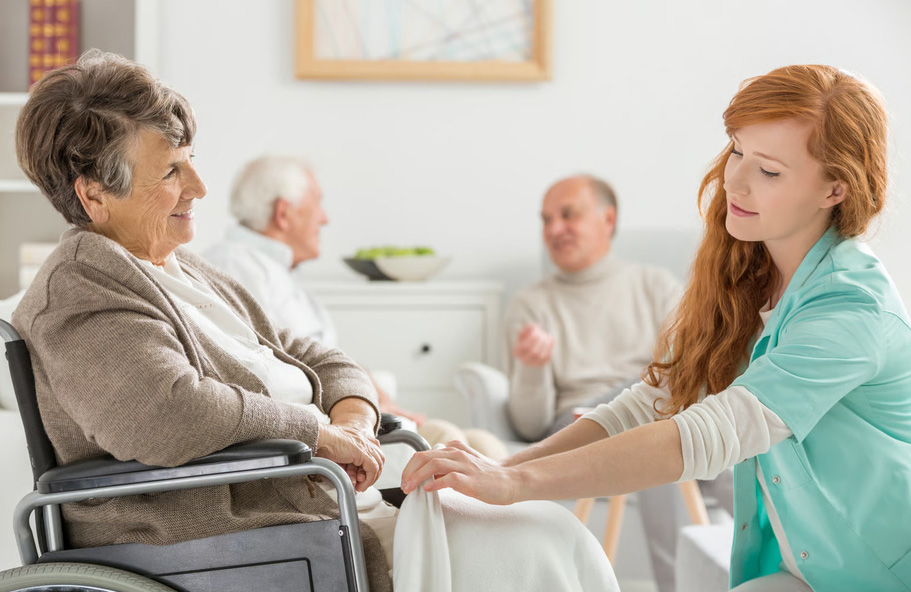 A caring female nurse in teal scrubs gently tending to an elderly woman in a wheelchair during a hospice care visit.