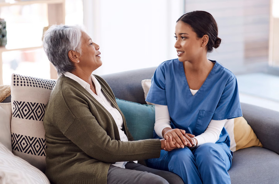 A young female nurse in blue scrubs sitting on a couch holding hands and talking with an elderly senior woman at home during a hospice consultation.